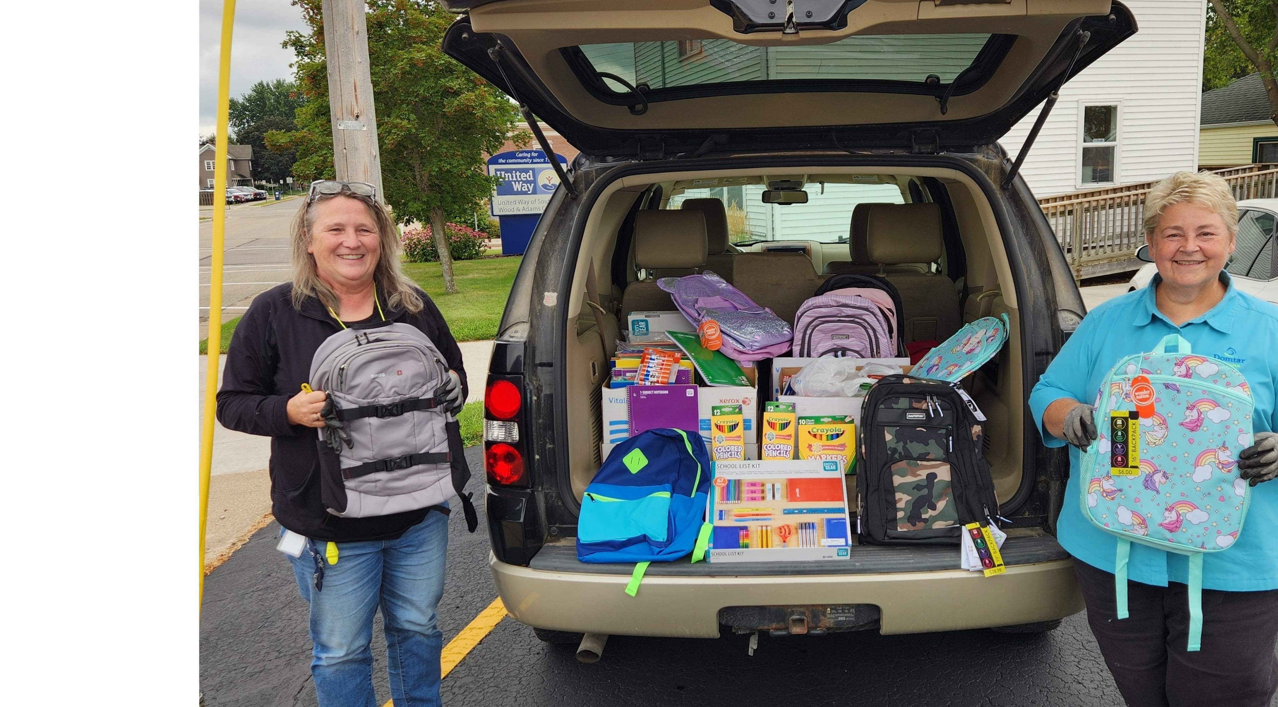 Two ladies holding backpacks in front of an open car trunk filled with school supplies.